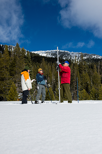 A researcher stands on a snow pack and measures how deep it is with a long measuring stick.