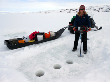 a photo of a smiling woman standing on ice and drilling holes in it