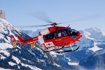 A photo of a red helicopter in the air over snowy mountains.