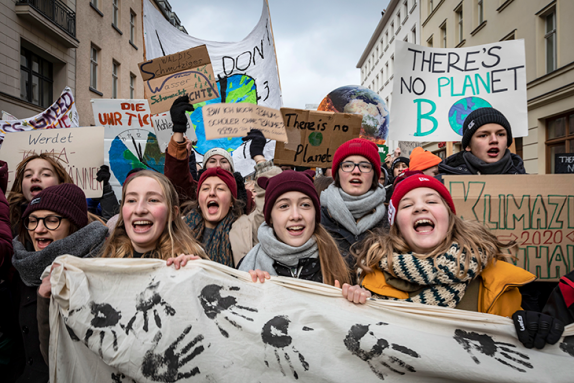 student marchers in Berlin Germany showing a variety of signs. In front is a row of students holding a banner with black paint handprints.