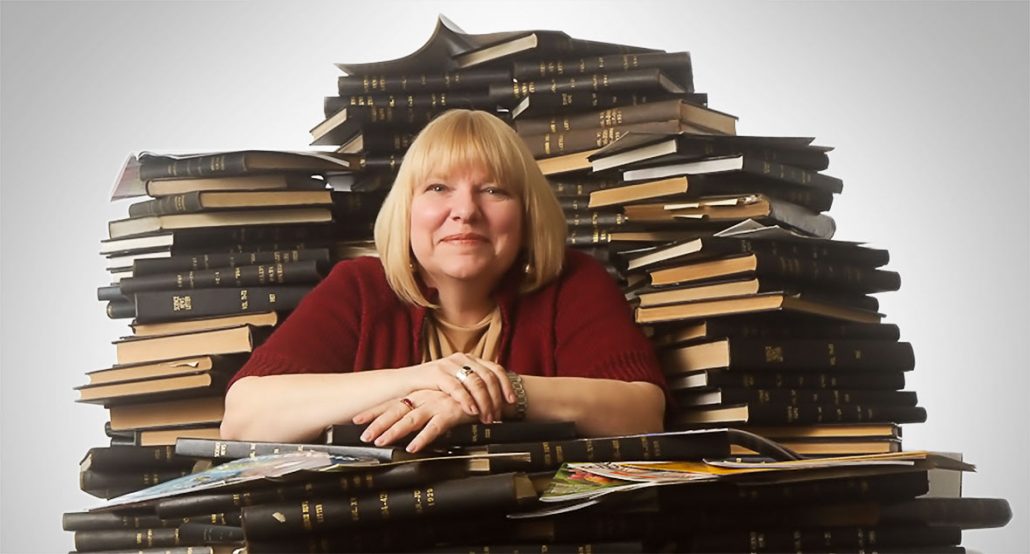 a photo of Janet Raloff sitting in front of piles and piles of old Science News magazines bound into books. Her hands are clasped on top of more books pile in front of her. She has blonde hair, a bob haircut and a big smile on her face.