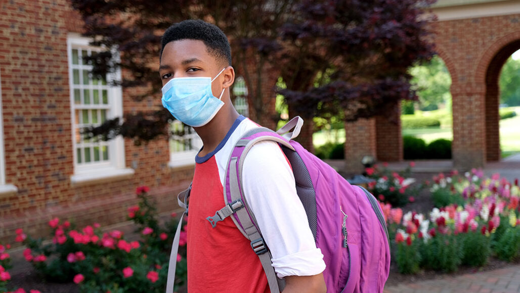 a photo of a young Black teen wearing a mask, he has a purple backpack on and is outside a school