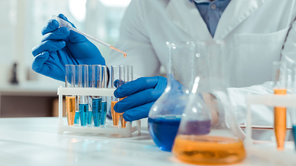 a photo of a table with orange and blue chemicals in test tubes and a gloved hand with a pipette adding more chemicals to the test tubes