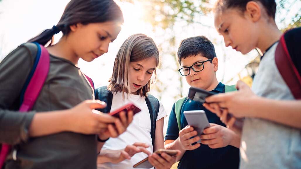 four kids wearing backpacks tand together in a group holding smartphones