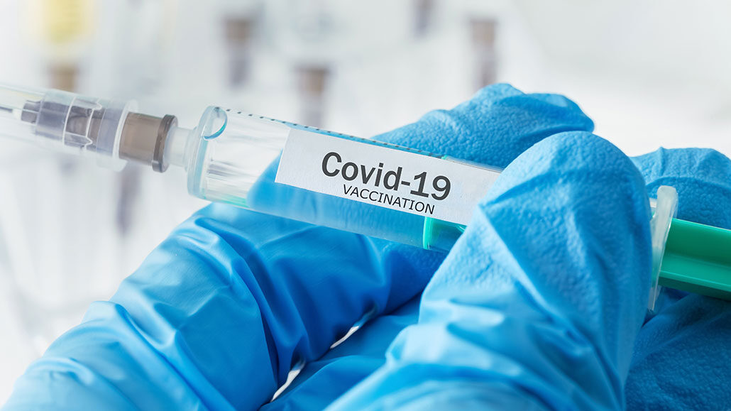 a photo of blue gloved hands holding a syringe labeled "Covid-19 Vaccination"
