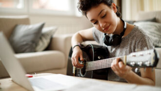 a photo of a girl playing a guitar in front of her laptop, she is wearing headphones around her neck