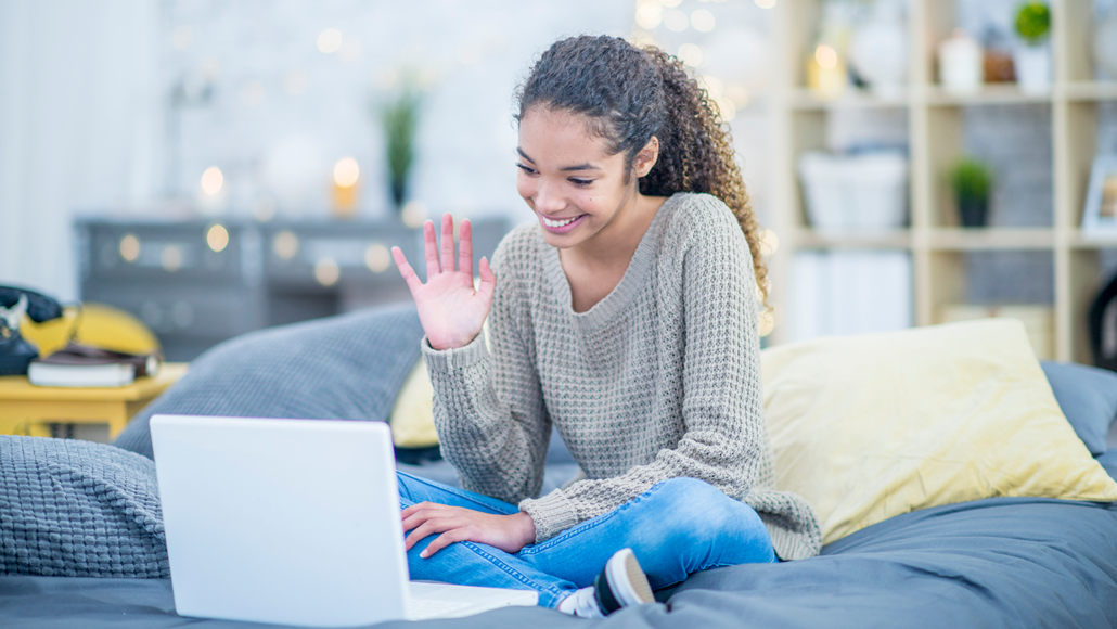 a teen girl with long curly hair waves at her computer screen while chatting online