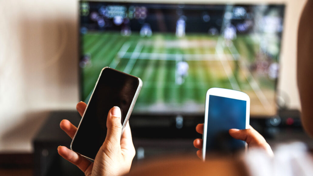 hands holding phones up in front of a TV showing a tennis match