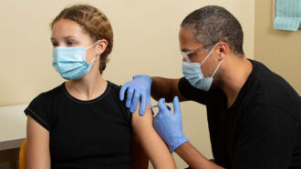 a Black girl looks away from the Black nurse giving her a shot in her arm
