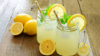 a photo of two jars of lemonade with straws on a wood table, cut lemons surround the jars