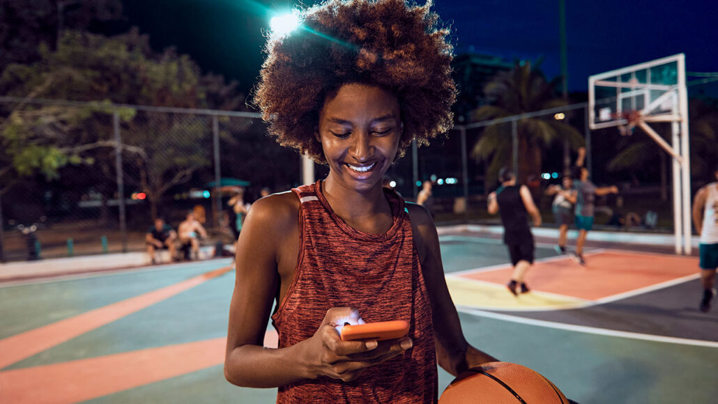 a young person with dark skin and an afro is standing on a basketball court at night and checking their phone