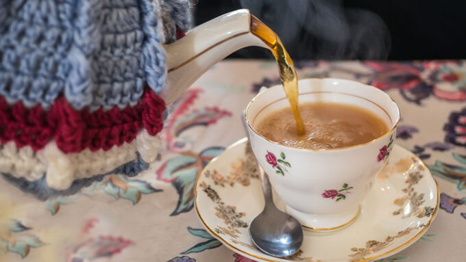 a teacup with a floral pattern is being filled with tea from a teapot covered in a crocheted tea cozy