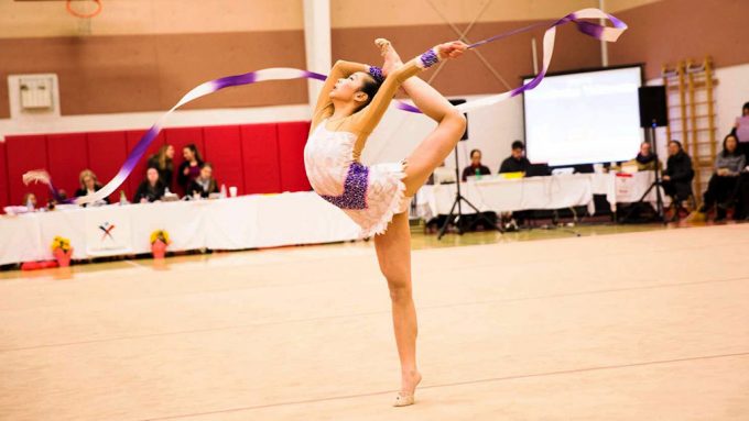 a photo of Michelle Hua wearing a sparkly rythmic gymnastics outfit in an indoor gym during competition. She is standing on one leg with the other leg arched and swirling a long white and purple ribbon