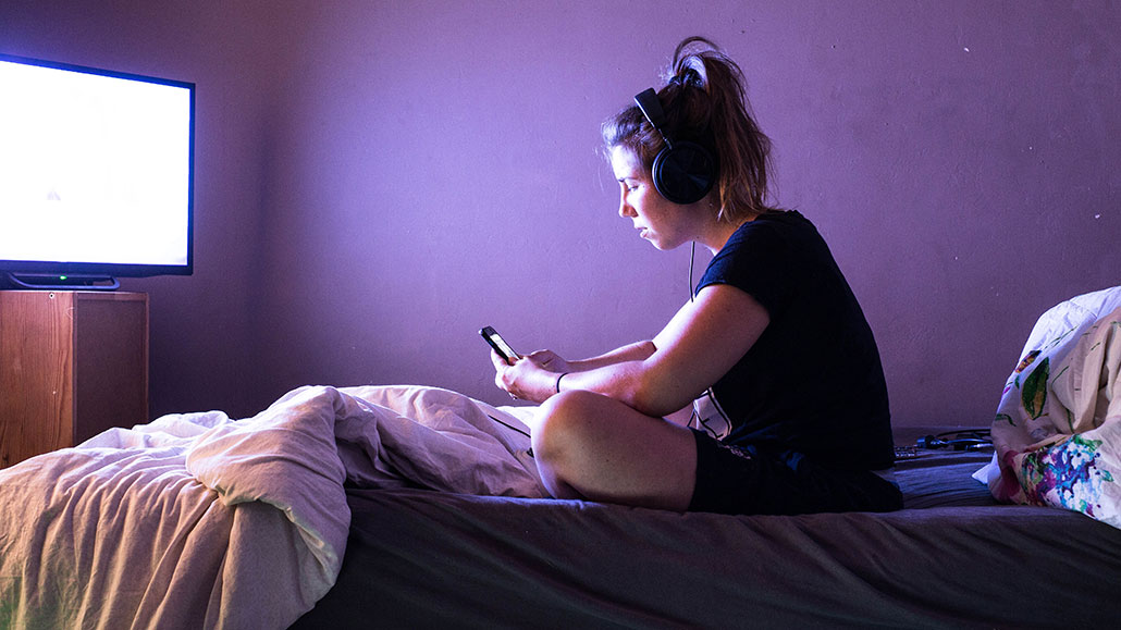 a woman looking at a cell phone on a bed in front of a large screen on a dresser. the screen is on and bright white