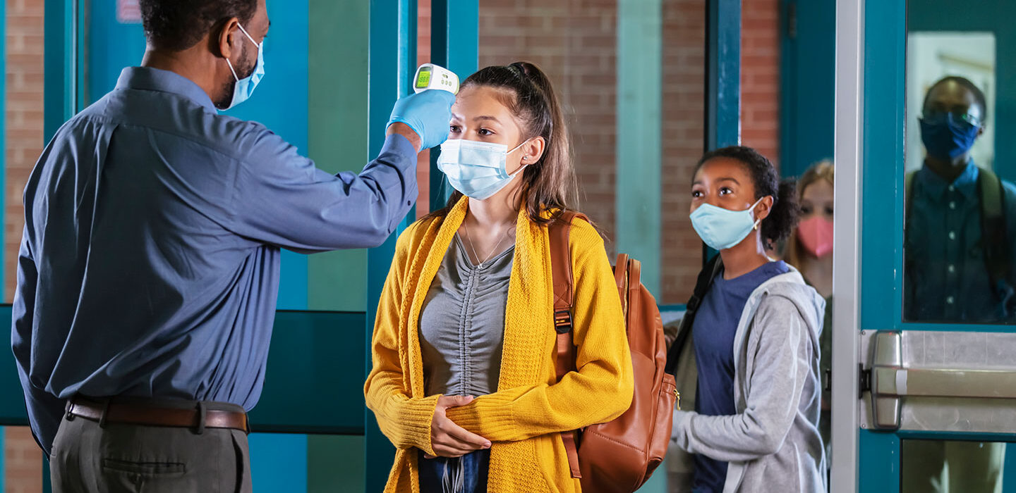 a young woman wearing a mask, yellow cardigan and a backpack getting her temperature taken as she enters a school