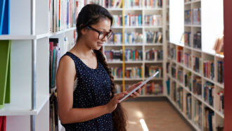 a female student looking at a tablet while standing near bookshelves