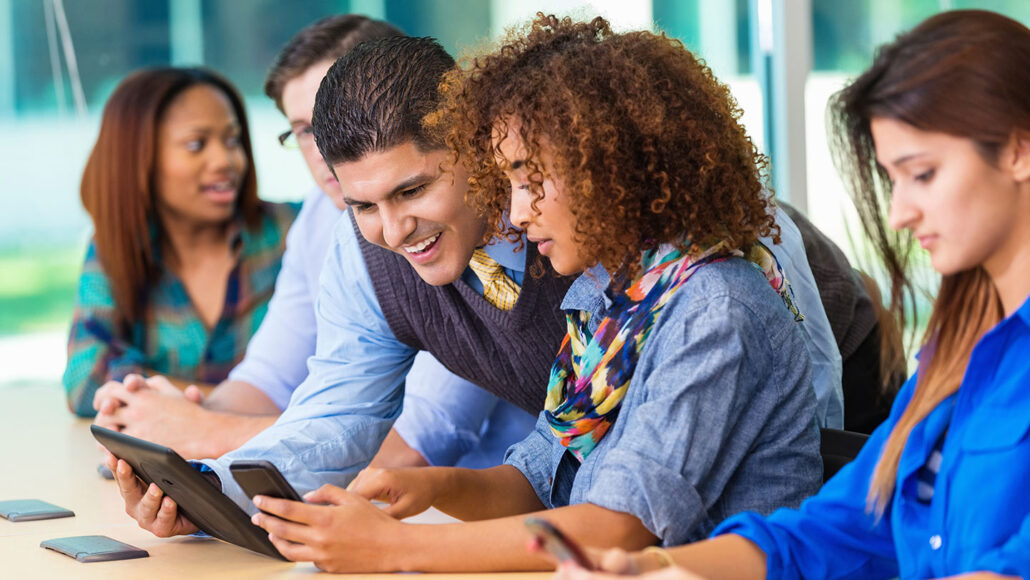 several students sitting at a desk looking at phones, tablets, and speaking with a teacher