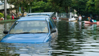 a car is partially submerged in a flooded street while people paddle in boats in the background