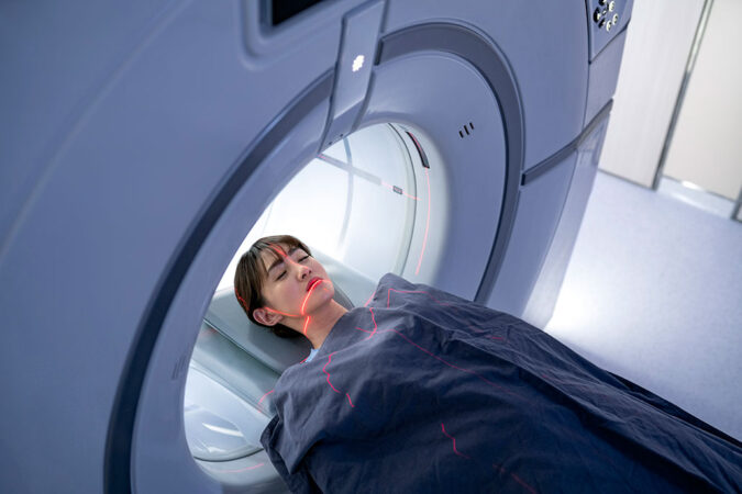 a woman lies on a mRI bed, a blanket is covering her up to her neck, her head is inside the machine