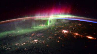 a view from the space station looking down on the night side of Earth with green and pink auroras rippling through the air high above ground