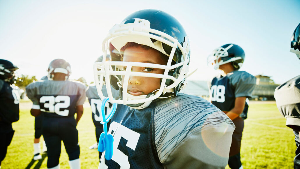 a Black teen football player faces the camera wearing his helmet and pads