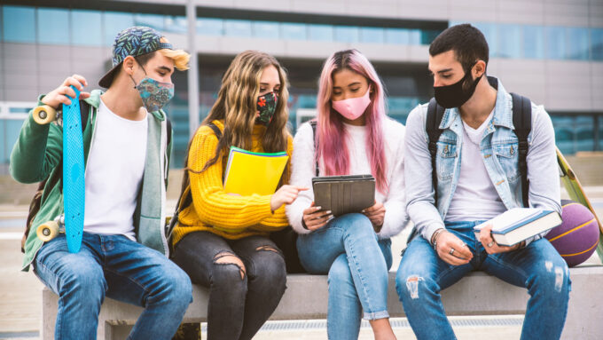 two teen boys and two teen girls sit together on a bench outside school, all wearing masks