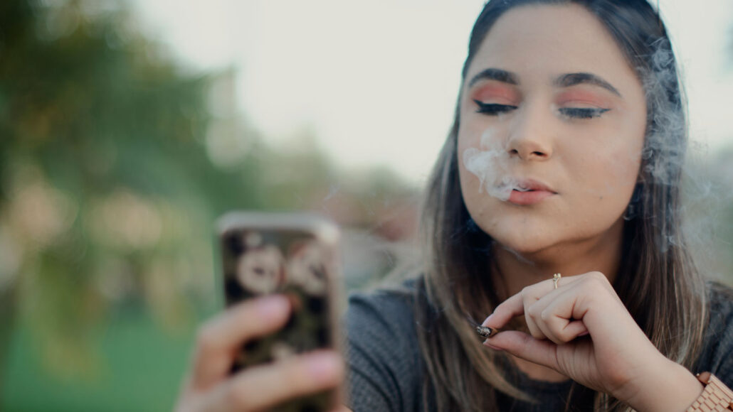 a teen girl smokes a joint while looking at her phone