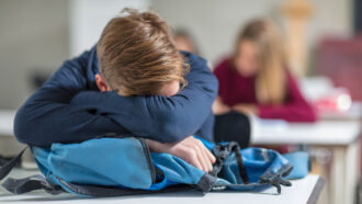 A boy rests his head on his backpack atop his desk to sleep in class
