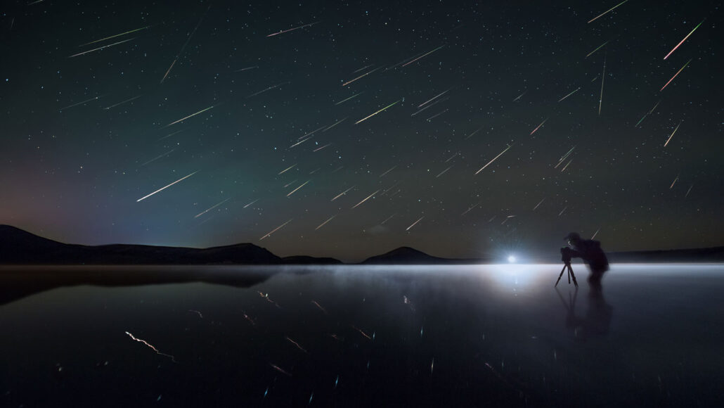 a dark landscape with the silhouette of a skywatcher, the sky overhead full of streaks of light that are meteors in the Perseid meteor shower