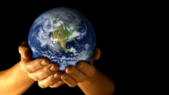 a pair of hands holds a model of the Earth against a black backdrop