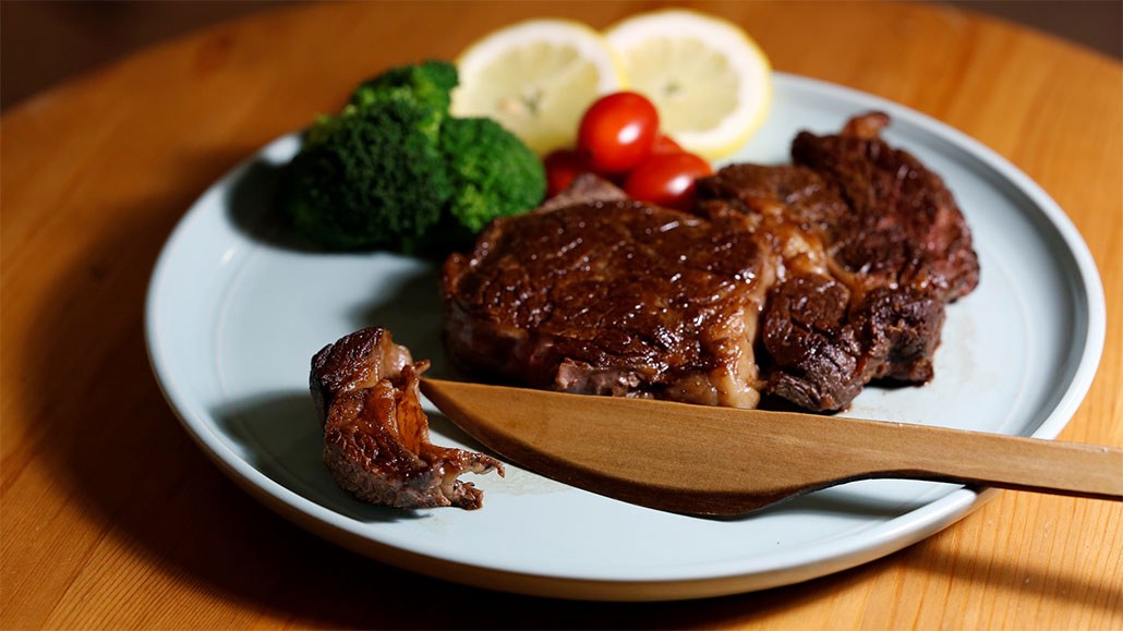 a photo of a dinner plate with a wooden knife, a steak with a small piece cut off of it, broccoli, tomatoes, and lemon