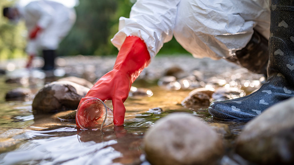 a person wearing gloves and hazardous material gear takes a water sample