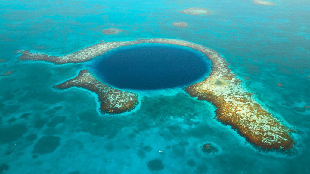 an areial view of a a ring-shaped coral reef surrounds the dark blue water of a lagoon in the lighter blue water of the open ocean