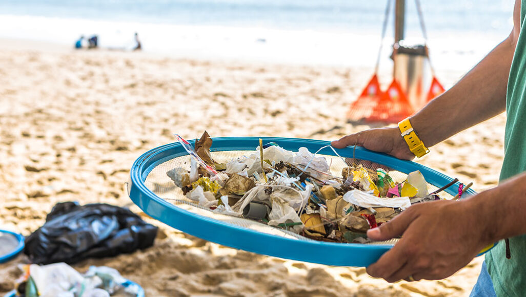 a photo of a beach. in the foreground are hands holding a blue mesh strainer full of plastic debris from the ocean