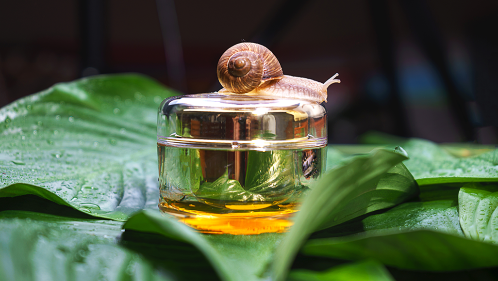 a photo of a shiny gold cosmetic container sitting on green leaves, a snail on top of the gold container