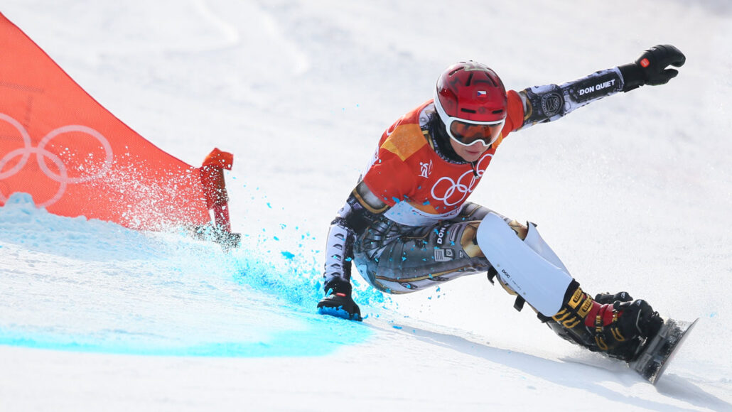 A snowboarder wearing the Olympic rings signal on her gear pulls a tight turn, leaning back to brush one hand agains the snow as she goes