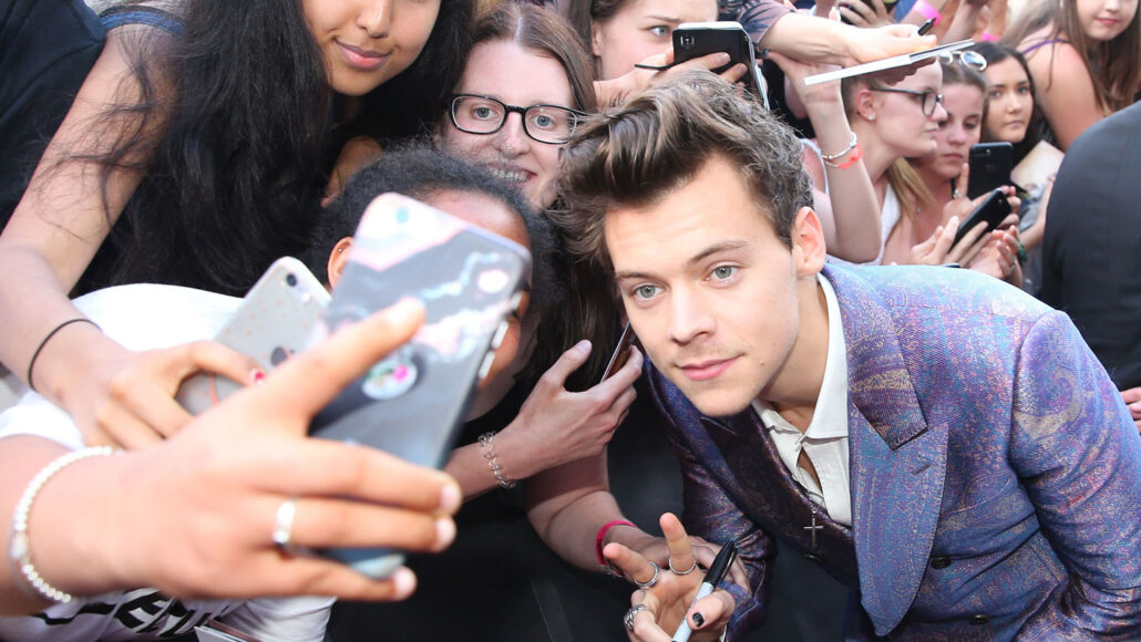 Harry styles, wearing a purple suit, poses for selfies with a crowd of fans