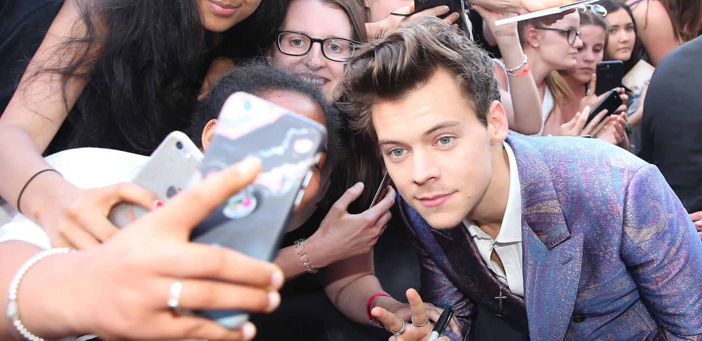 Harry styles, wearing a purple suit, poses for selfies with a crowd of fans