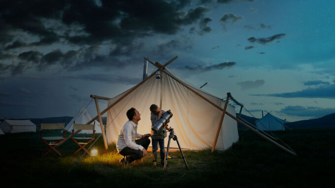 A father kneels next to a child in front of a tent as the kid looks through a telescope at the night sky