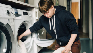 a genderfluid teen wearing a black hoodie is loading a washing machine