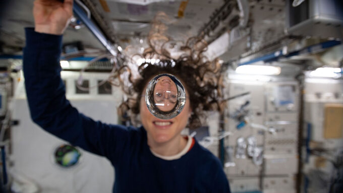 A female astronaut with curly hair on the International Space Station. She is looking at the camera through a floating gob of water, through which her face appears upside down.