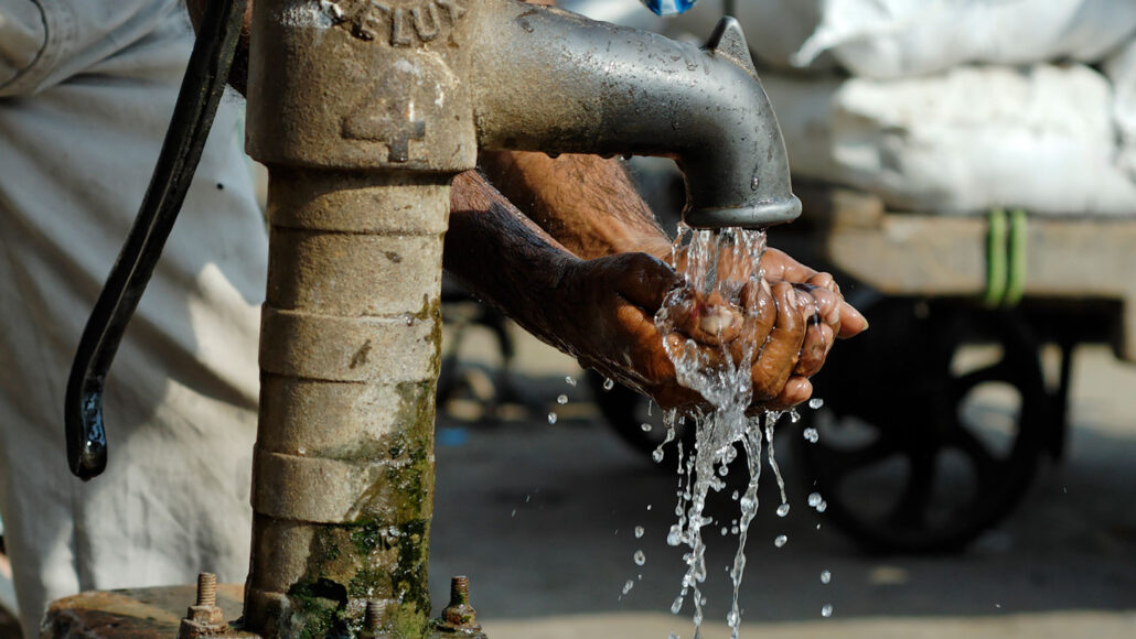 a pair of hands are cupped beneath the faucet of a borehole in the ground