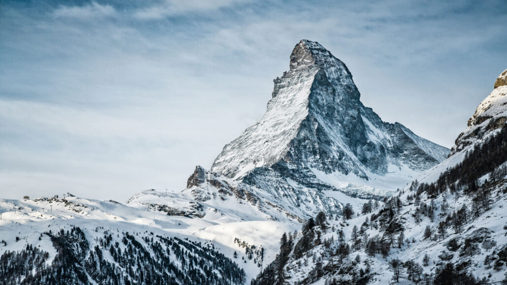 a snow-dusted mountain peak in a range stands out against a cloudy blue sky