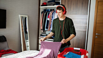a teen boy wearing headphones folds laundry from a basket on his bed