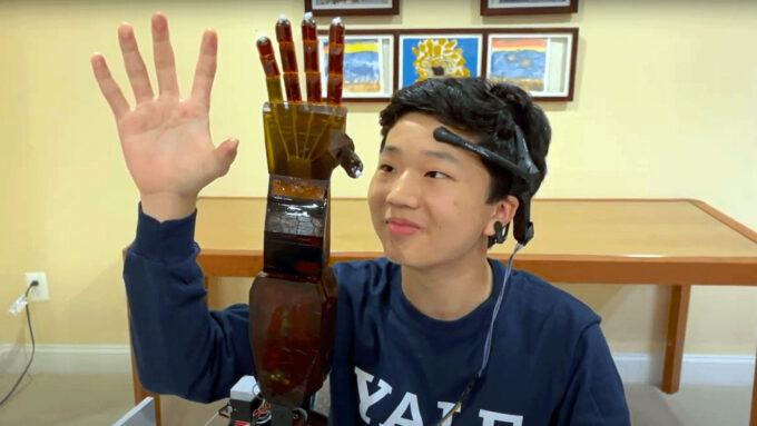 a teenage boy holds his hand up, palm open, beside a robotic arm in the same position, while he wears a headset on his forehead