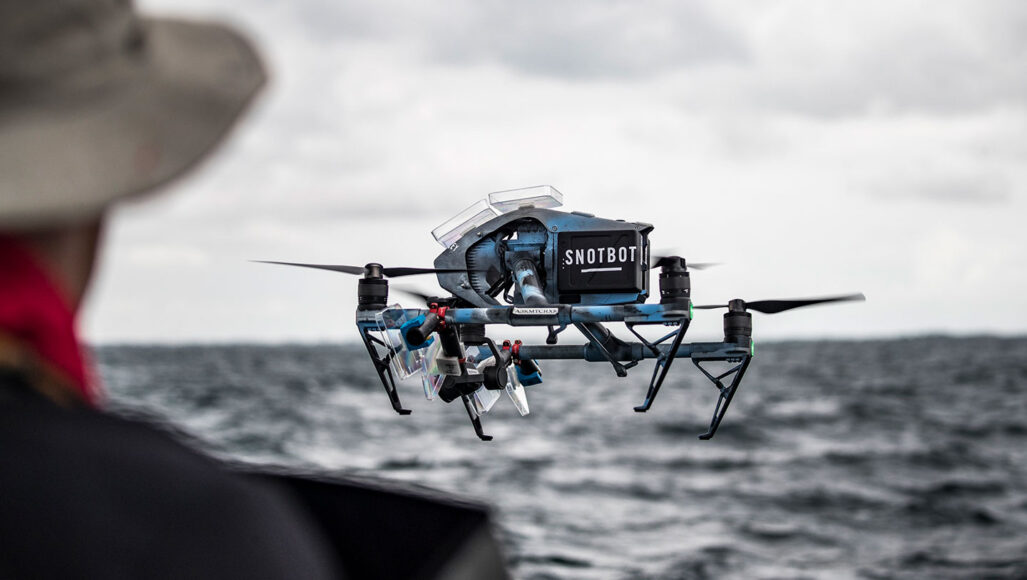 a photo of a blue drone hovering in the air. The drone has a sticker that reads "SnotBot" seen over the shoulder of a man partially in the left most edge of the image.