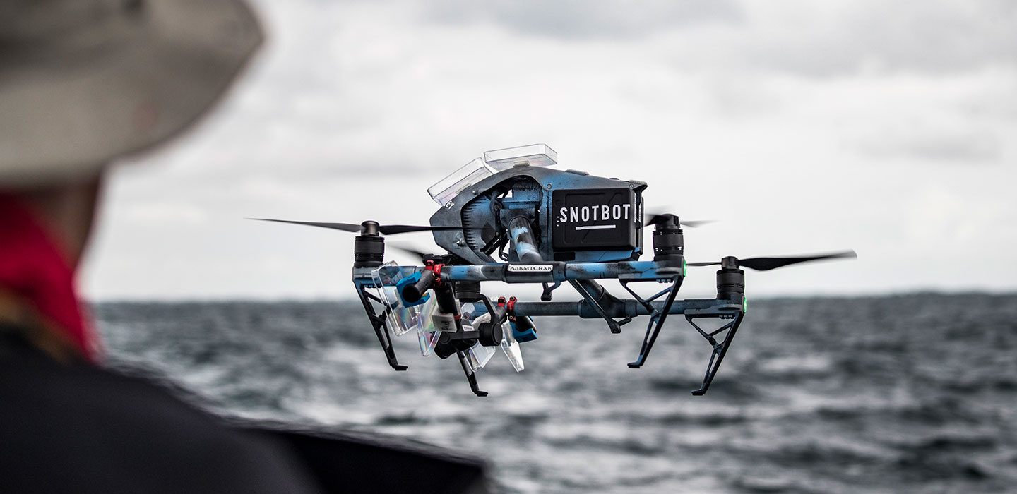 a photo of a blue drone hovering in the air. The drone has a sticker that reads "SnotBot" seen over the shoulder of a man partially in the left most edge of the image.