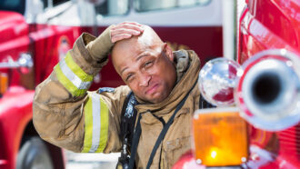 a photo of a fireman dressed in fire gear. He is sitting on a fire engine and sweating profusely. He looks miserable and very hot.