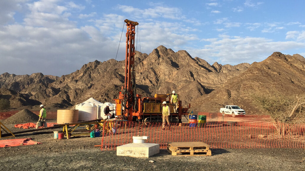 a large orange drill is in center of the picture. there are workers around the drilling site and mountains in the distance at the horizon.