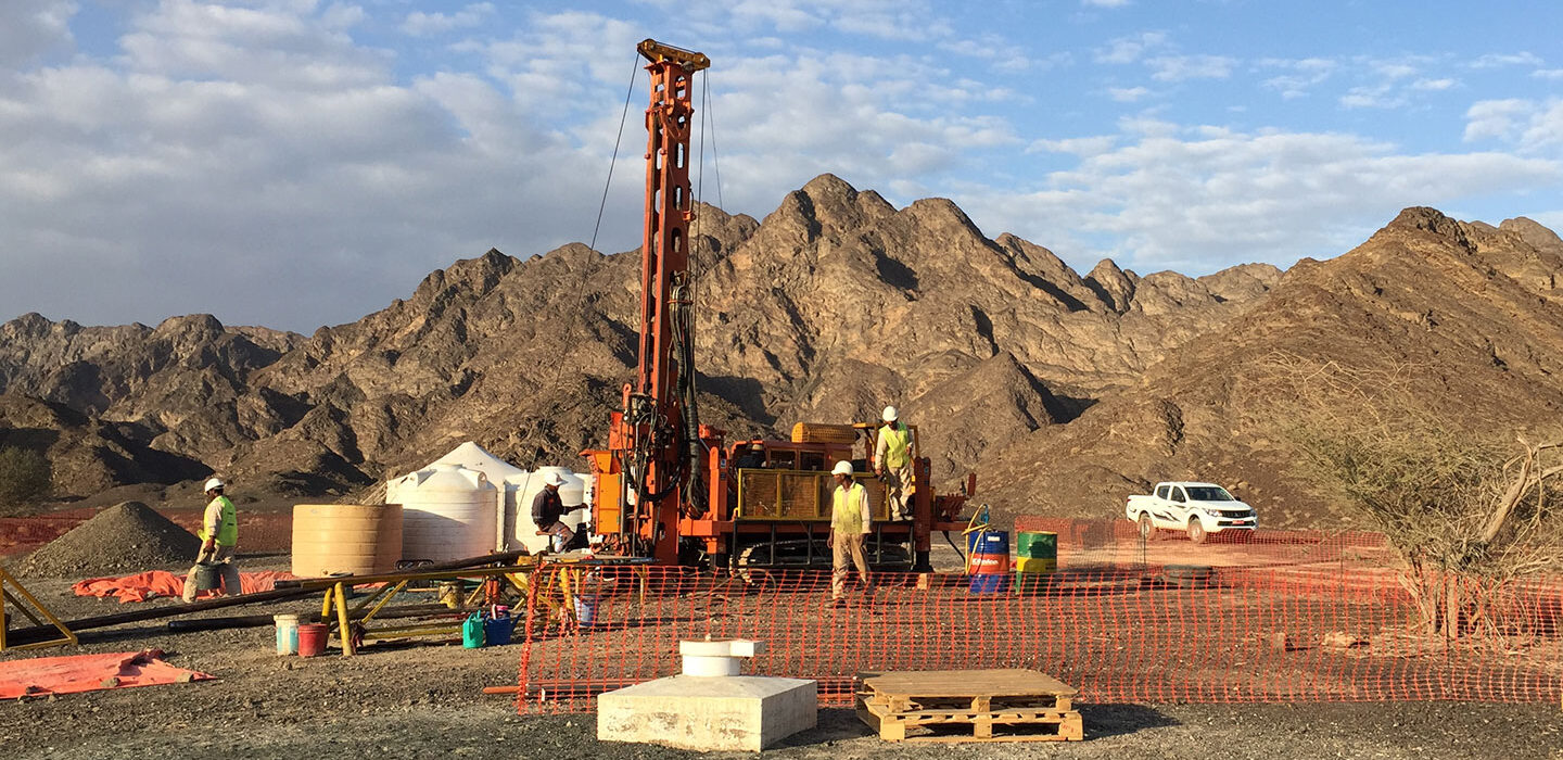 a large orange drill is in center of the picture. there are workers around the drilling site and mountains in the distance at the horizon.